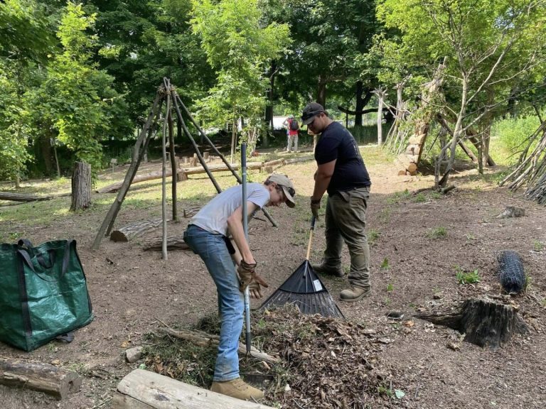 Two people raking leaves next to a rustic wooden structure