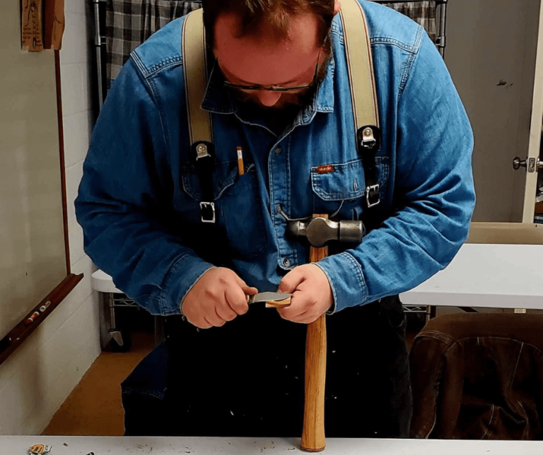 A man in suspenders whittling and holding a hammer