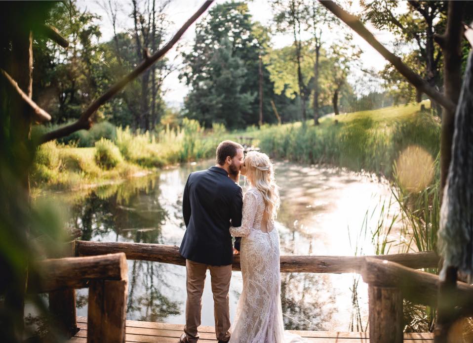 Bride and groom embracing on a wooden dock beside a pond at golden hour, framed by tree branches.