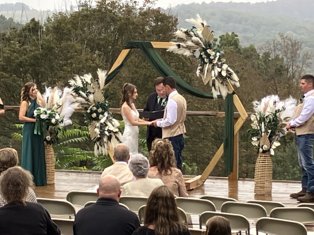 Bride and groom exchanging vows at a floral arch while an officiant and seated guests look on, mountains in the background.