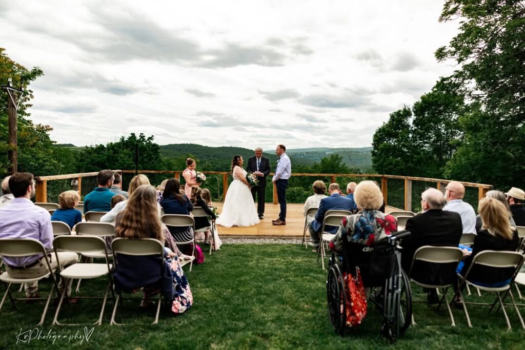 Outdoor wedding ceremony with officiant and couple at the altar, guests seated in folding chairs, mountain view in the background.