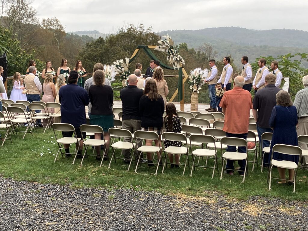 Wedding ceremony viewed from behind rows of seated guests, facing the floral arch and mountain backdrop.